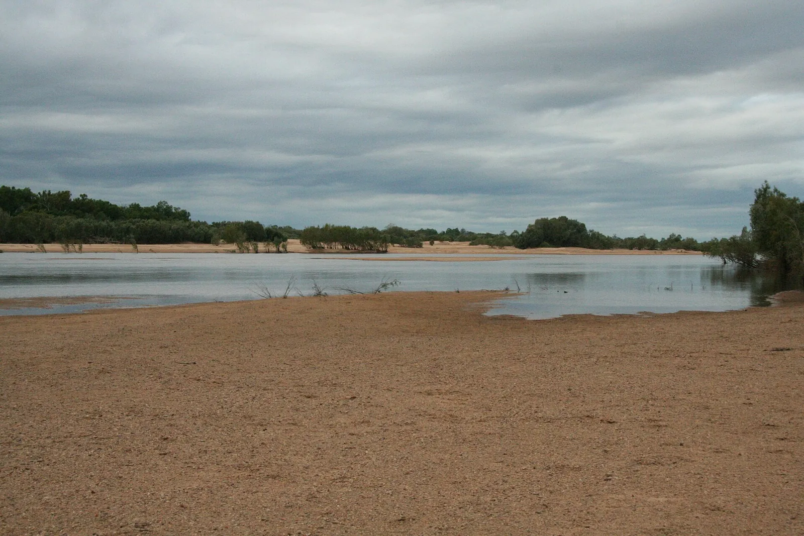 A wide sandy river bed with a shallow channel of water and trees on the far bank, in the dry season